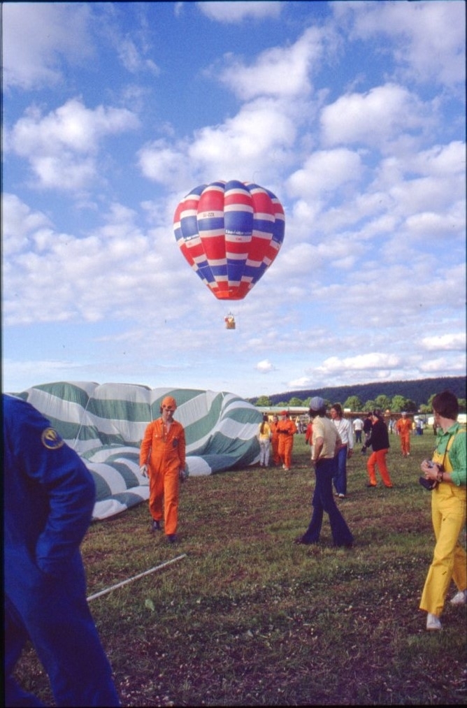 En röd-blå-vit ballong i luften. I förgrunden personer i orangea overaller vid ett grön-vit randigt ballonghölje som ligger på gräset.