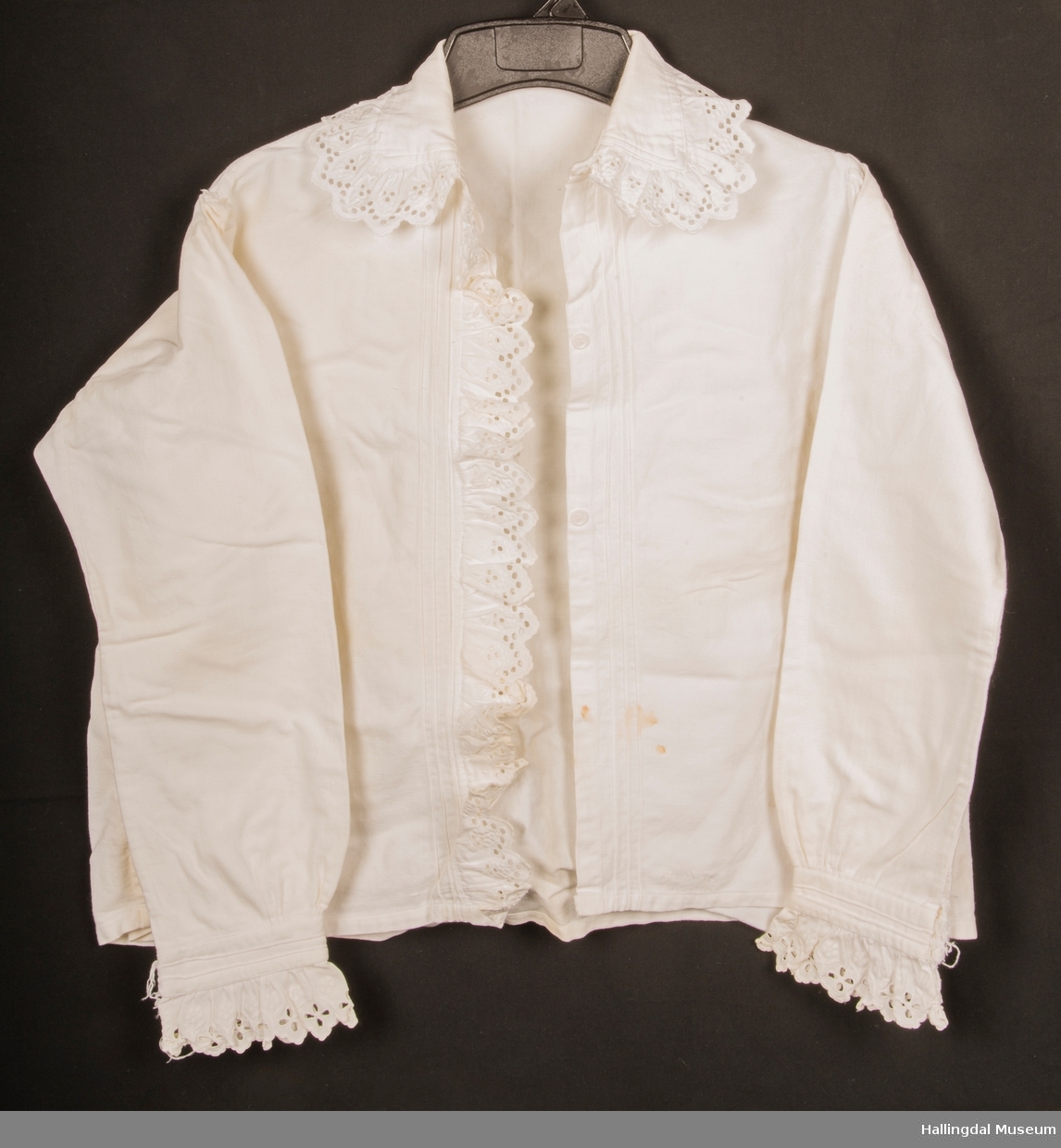 Hjemmelaget bluse sydd av bomullstøy med blonder rundt kragen, håndledd og langs åpningen. Skjorta er lukket med 3 plastknapper som er skjult bak blonden når skjorta er lukket.