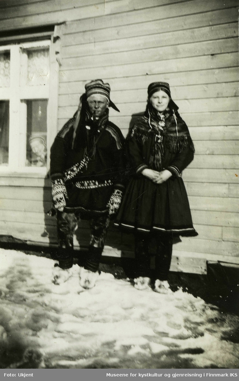 Portrett av en mann og en kvinne i samiske klær. De står foran et trehus. Til venstre er et vindu og man ser at en kvinne med samisk lue følger fotograferinga. Det ligger sne i bakken. Sted, personer og fotograf er ukjent.