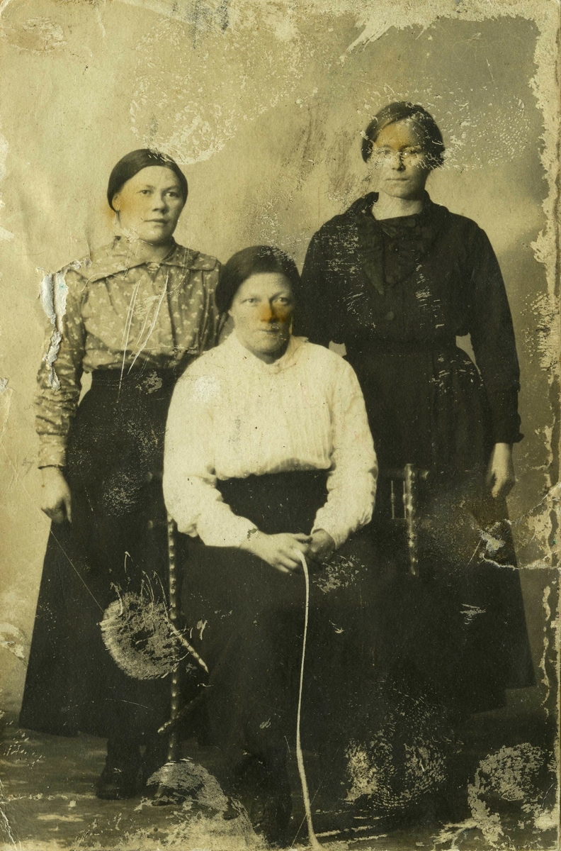 Studioportrett av tre kvinner. Kvinnen i midten sitter på en dreied trestol. De to andre står bak henne. Alle tre har på seg skjørt og bluser. De avbildete personer og fotografen er ukjent.