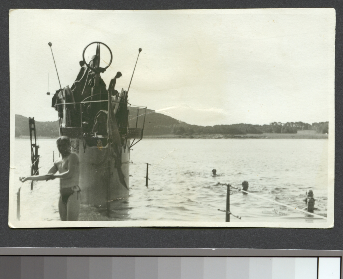 Bilden visar manskapet från ubåten Najad som tar en simtur runt båten som har dykt ner så mycket att bara tornet sticker ovanför vattenytan. I förgrunden står en man i badkläder.