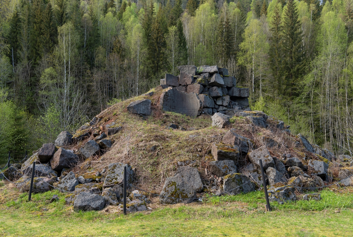 Ruinen etter Fritzøe jernverks masovn på Moholt i Siljan. Fotografiet er tatt fra sør. Det rennende elvevatnet som drev blåsebelgene som tilførte den metallurgiske prosessen som foregikk her nok oksygenrik luft til at temperaturen inne i ildstedet ble høy nok, er her skjult bak ruinen.

Masovnteknologien ble innført i Norge tidlig på 1600-tallet. Det dreide seg om digre, høye steinovner som hadde noenlunde runde brennkamre. Når en slik ovn skulle settes i drift, ble den fylt med trekol, som ble antent og fikk stå å gløde 3-4 uker før konstruksjonen var varm nok. Tilstrekkelig høy varme ble oppnådd ved hjelp av oksygentilførsel fra et par blåsebelger som ble drevet ved hjelp av vannkraft fra elva.  Etter at ovnen hadde stått full av glødende trekull et par uker, var murverket varmt nok til at den metallurgiske produksjonsprosessen kunne starte. Det ble gjort ved «påsettinga» av trekol og malm, lag for lag, fra «krona», overkanten på masovnmuren. Slik kunne masovnen holdes i kontinuerlig drift gjennom et par år før det ble nødvendig å avslutte fyringa og skifte ut steinene i de mest varmeeksponerte delene av brennkammeret. Dette forutsatte naturligvis at verket hadde gode lagere av malm og trekol. Mens prosessen pågikk ble det stadig tappet råjern gjennom et hull - «stellet» - i den nedre delen av ildstedet. Dette jernet kunne enten brukes i støpejernsprodukter umiddelbart, eller det kunne bearbeides videre til smidigere smijern i en «hammer».  På Moholt ble det bygd et hammerverk tidlig i 1730-åra. Hit transpporterte man forholdsvis små volumer av råjern fra Fritzøe-verkets hovedanlegg i Larvik med ferger over innsjøen Farris, i stedet for å frakte store volumer av trekull den motsatte vegen. Under napoleonskrigene tidlig på 1800-tallet økte etterspørselen etter jern. I denne perioden hadde det vist seg vanskelig å skaffe nok trekull til masovnen i Barkevik i Brunlanes. Derfor bestemte ledelsen i Fritzøe-verket at noe av råjernproduksjonen skulle flyttes til Moholt, der man hadde bedre tilgang på trekull fra skogene i Siljan. Dette var bakgrunnen for at man bygde den masovnen vi ser på dette fotografiet i perioden 1809-1817. Årsaken til at byggeprosessen tok så lang tid var at de varmeresistente steinene som skulle være i området rundt stellet måtte importeres, noe som var vanskelig i en periode med krig og blokader av skipsfarten. Masovnen på Moholt var i drift i en 50-årsperiode, fra 1817 til 1867. Den var omgitt av ei smeltehytte, en bordkledd bindingsverkskonstruksjon, som vi ser noen av fundamentene til i forgrunnen på dette bildet. Dette kulturminnet er tilgjengeliggjort for interesserte med informasjonsskilt og tilstøtende rasteplass. Klatring i ovnsruien er ikke ønskelig. Den er derfor skjermet ved hjelp av et enkelt kjettingstengsel. 

Litt mer historisk informasjon om den metallurgiske virksomheten på Moholt finnes under fanen «Opplysninger».