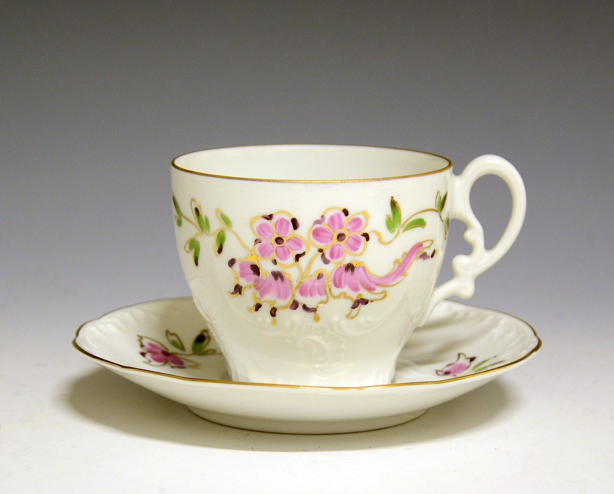 Kaffeskål av porselen. Fanen dekorert med rosa blomster og grønne blader. Gullstrek rundt kanten.
Modell: 332.4