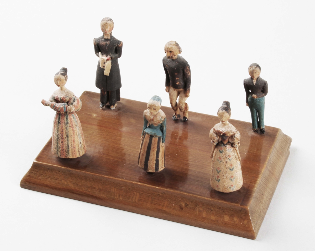 Figurgrupp i trä. "Symboliserar ett dop". 1830-tal. 6 stående figurer, i snidat trä, med handmålad dekor. I form av, präst, kvinna med barn i famnen, 2 kvinnor och 2 män. De sitter med stift, i rektangulär, brunbetsad- och lackad träplatta, med insvängda kanter.

Till denna figurgrupp hör konsolhylla BM 39798.