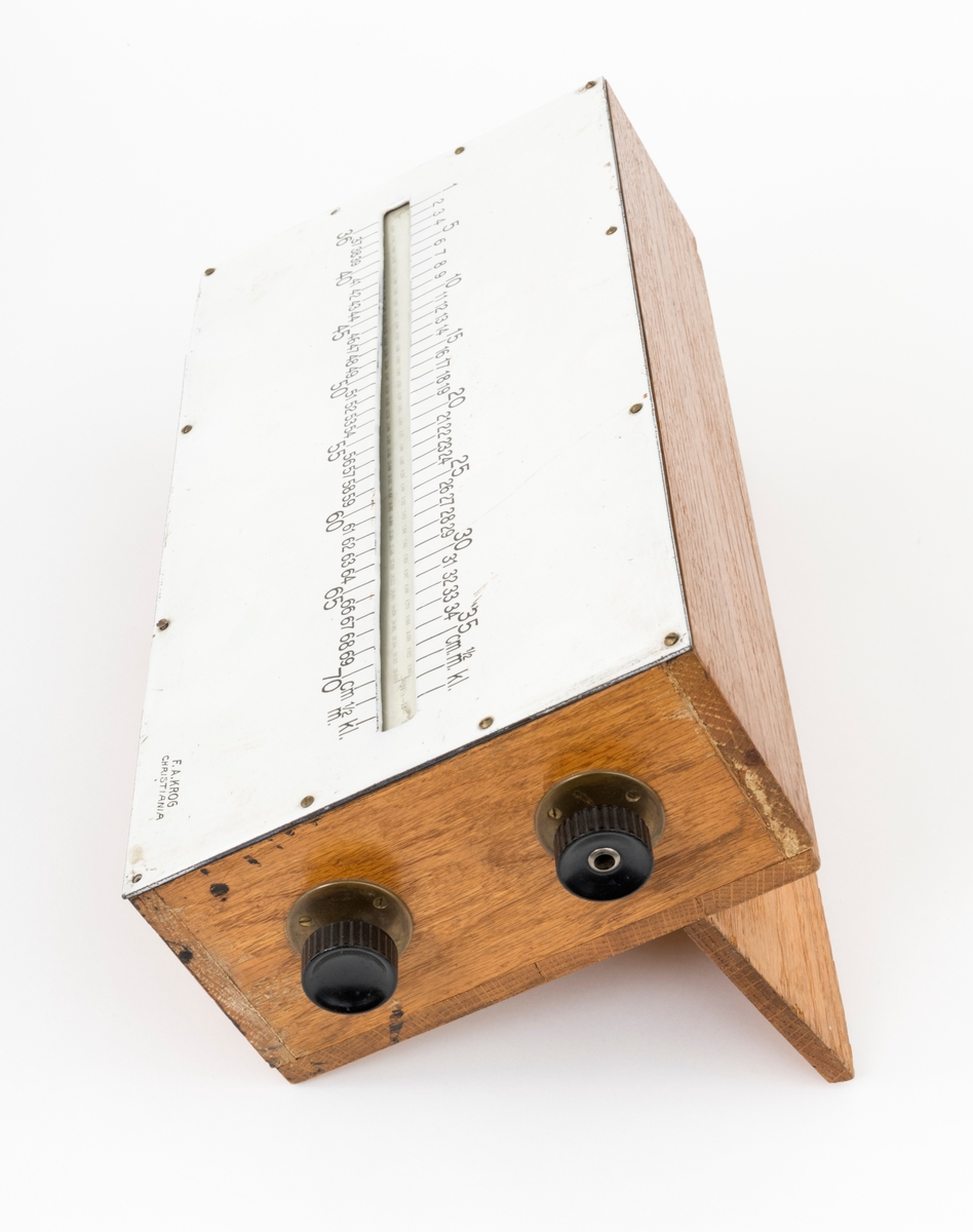 Kubikkberegningsapparat, levert av produsenten F. A. Krog i Skovveien i Kristiania, antakelig tidlig på 1900-tallet.  Apparatet er formet som et rektangulært skrin, utført i eiketre, men med «lokk» av ei emaljert jernplate.  Langs midtaksen på dette lokket er det ei lang, smal, rektanuglær spalte med glass under.  Gjennom denne ser vi ned på en tabell.  Tabellen er trykket på en papirrull, som er forankret på to valser nede i skrinet.  Tabellen kan beveges ved hjelp av dreiehjul på skrinets endeflater, to på hver side, ett i hver ende av de nevnte valsene.  Lengst til høyre i tabellvinduet finner vi «klassifiseringsinformasjon» - angivelser av stokklengder, diametere og salgsklasser.  Langs den øvrige delen av vinduet kan man lese av kubikkmeterangivelser for fra 1 til 70 stokker med samme dimensjoner og klassespesifikasjon.  Avlesinga skjer fra en tolinjet tabell på den nevnte papirrullen inne i skrinet, og med stokketallesangivelser på lokket - 1-35 stokker på oversida av vinduet og 36-70 stokker på undersida.  Det nevnte emaljerte lokket, som er hvitt med svarte tall og bokstaver, er skrudd fast til toppen av skrinet.  Det er grunn til å tro at valsene med tabellen må være montert før dette ble påsatt.  Skrinet er ellers lagd av høvlet eiketre, og den holdes sammen av de nevnte skruene i lokket og spiker fra botnstykket.  På standflata er det også hengslet en klaff, som kan vippes opp, slik at lokket skrår mot brukeren når apparatet brukes på et bord eller et annet plant underlag.

Apparatet er brukt ved utregning av tømmernotaer.