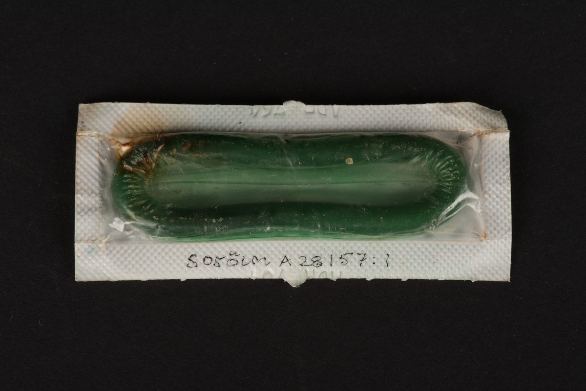 En kondom i förpackning. Kondomen är grön. Förpackningen är genomskinlig på ena sidan. Den andra sidan är vit med blommor i grönt.