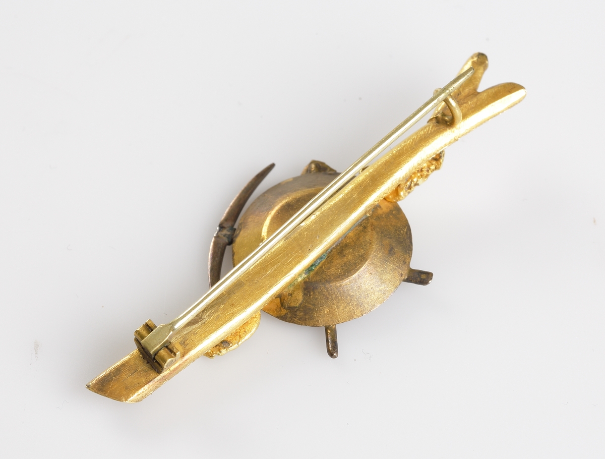 Guldgrävarnål som består av en trädgren med en guldvaskarpanna med texten "YUKON" i botten placerad ovanpå. Vaskpannan är fäst vid grenen med en guldnugget på vardera sidan. Över vaskpannan ligger en hacka och en spade korslagda.