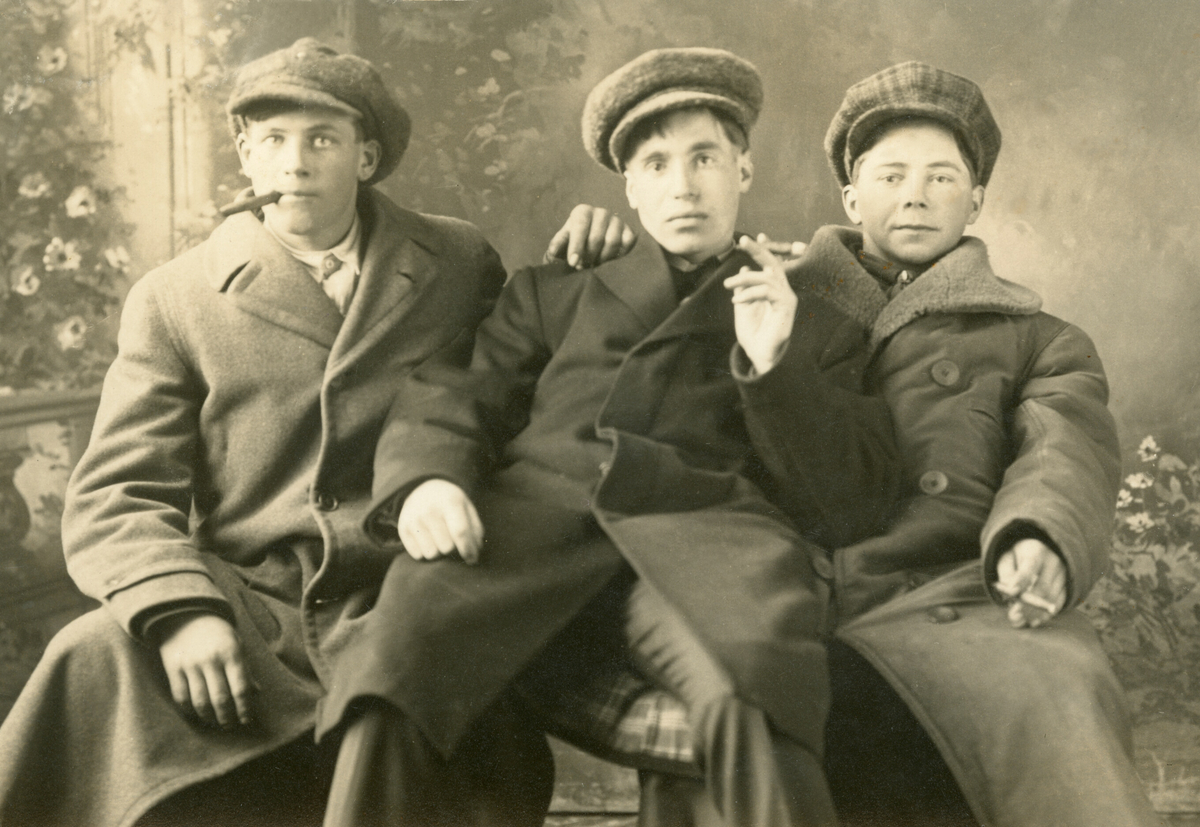 Tre yngre menn i frakkar og luver sit i fotoatelier og røyker.
Personane er ukjente.
Laga som prospektkort