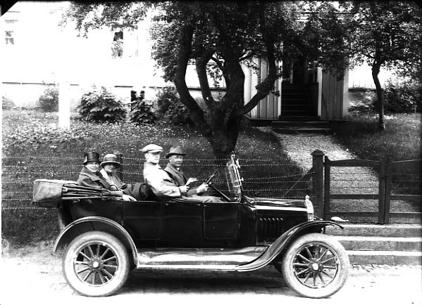 En bil (cabriolet) står parkerad på Brahegatan i Gränna. I framsätet sitter en äldre och en yngre man. I baksätet sitter två kvinnor.
Någon av männen är sannolikt Sven Larsson från Kronäng, Kinna