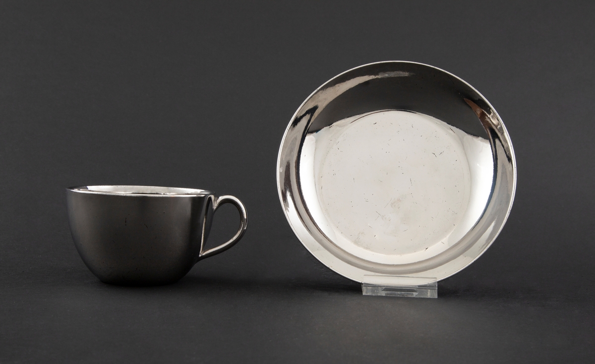 Kopp med skål i keramikk, glasert med sølvfarget luster. Tilnærmet sylinderformet korpus med påsatt hank. Lav sirkulær skål.