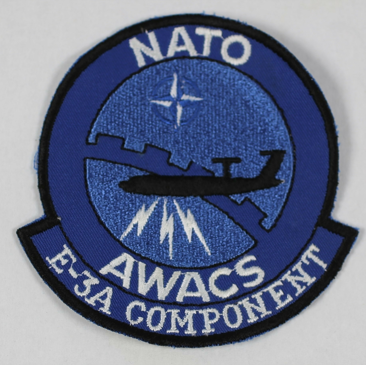 Sort silhuett av AWACS på blå bakgrunn med NATO stjerne,  og tre lyn
