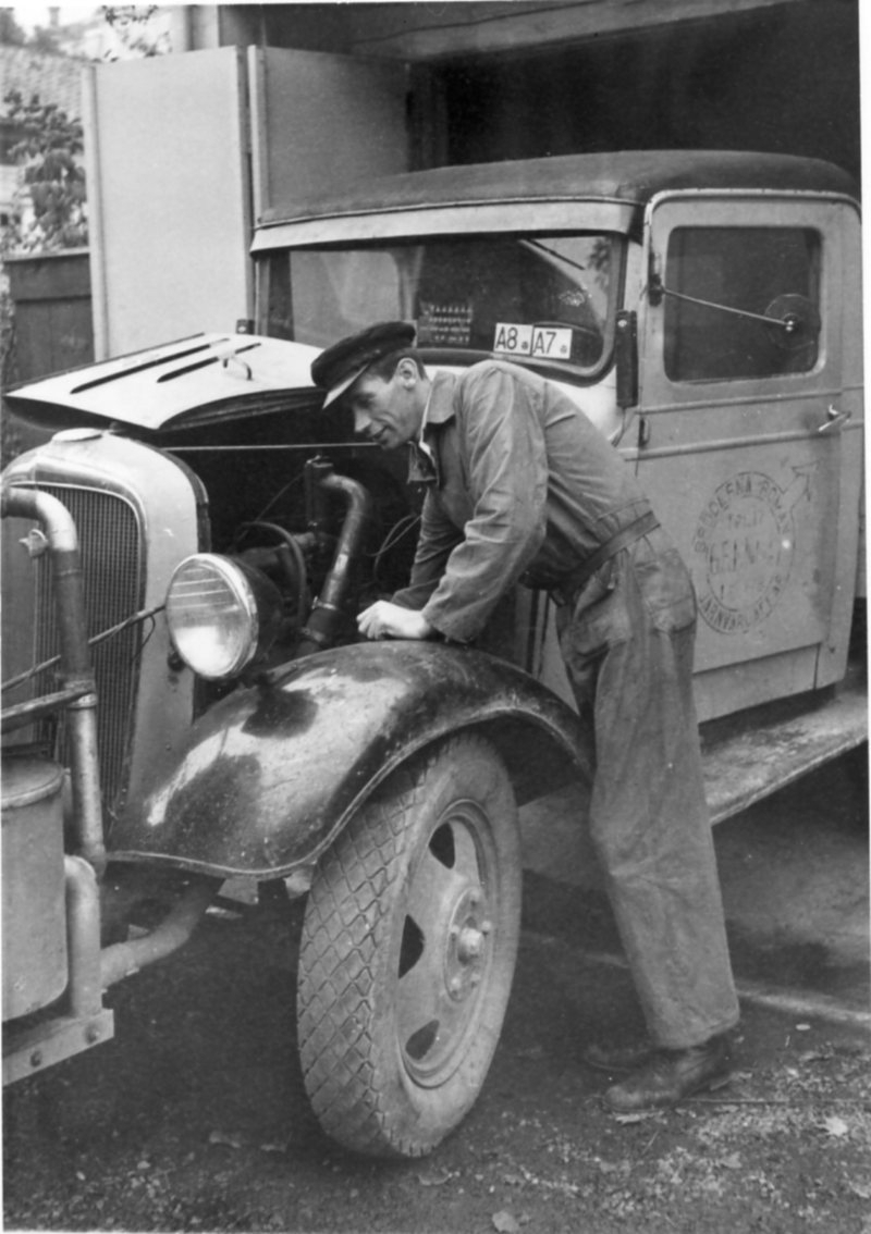 En man i overall och keps står lutad över motorn på en mindre lastbil som delvis står i ett garage. Dörren har en logo med texten: "Brödern Roman Järnvaruaffär".
Lastbilen är försedd med gengasaggregat.