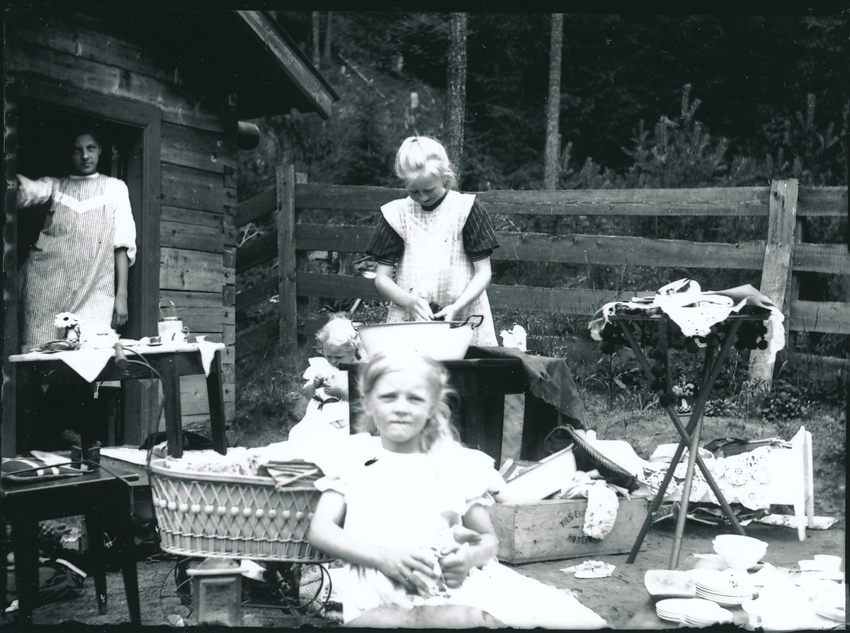 Tre småjenter i ferd med å vaske og leke utenfor hytte. En kvinne står i døråpningen. Utenfor hytta står bl.a. bord, stoler, klær og dukkevogn. Gjerde i bakgrunnen med skogen utenfor.