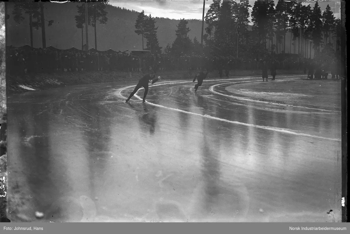 Juniormesterskapet på skøyter i 1932. To skøyteløpere under konkurranse. Folkemengde på tribunen.
