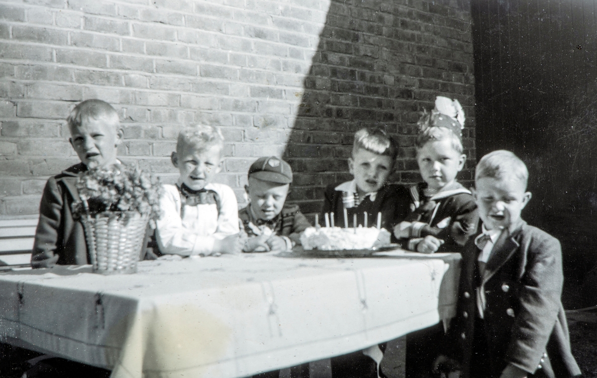 Bursdagselskap, barne bursdag. Arne Hesselberg, født 27.mai 1947, feirer sin 6-års dag (6 lys på kaka) sammen med kamerater.
Tatt inne i gårdsrommet til Torggata 113 mot den vestre ytterveggen til St. Torfinn kirke. 1953.