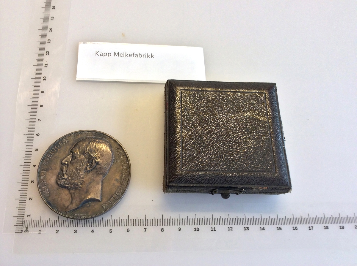 Etui med gråfarget mynt avbildet med Oscar II Norges konge på en side og en karakter fra gresk eller romersk mytologi på andre siden.