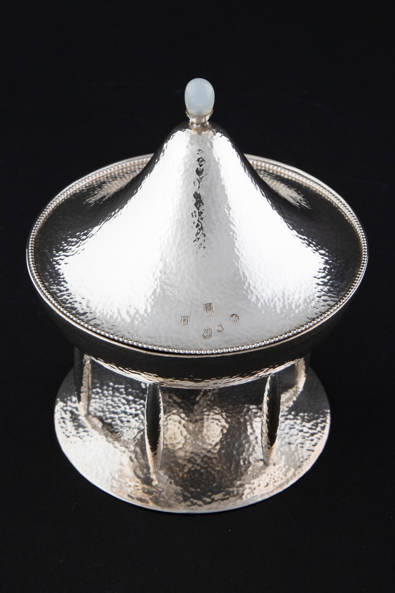 Bonbonnière med lokk i hamret sølv. Skålen er halvkuleformet og står på seks føtter festet til en sirkulær, svakt hevet fot. Lokket har konisk form og ender i en lokknapp med en opal. Langs kanten er lokket dekorert med perlestav.