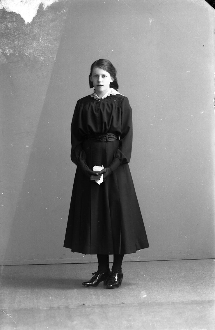 Porträtt av en flicka i mörk klänning. Hon håller en liten bok (psalmbok?) och en näsduk i händerna. Troligen konfirmationsbild.