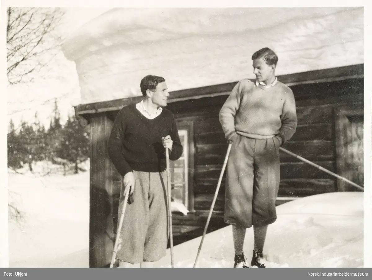 Solli februar 1931. To menn i nikkers ser på hverandre. De har ski på bena og skistaver i hendene. de står foran laftet hytte med mye snø på taket.