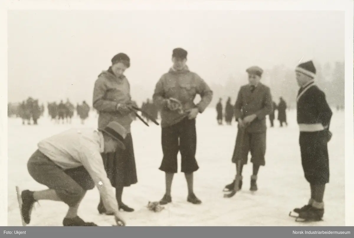 Fra Holtekilen, januar 1932. En  mann med skøyter på bena lener seg ned på isen. Fire andre, en kvinne og tre menn ser på han. Flere av personene har også på skøyter. I bakgrunn flere mennesker på isen.