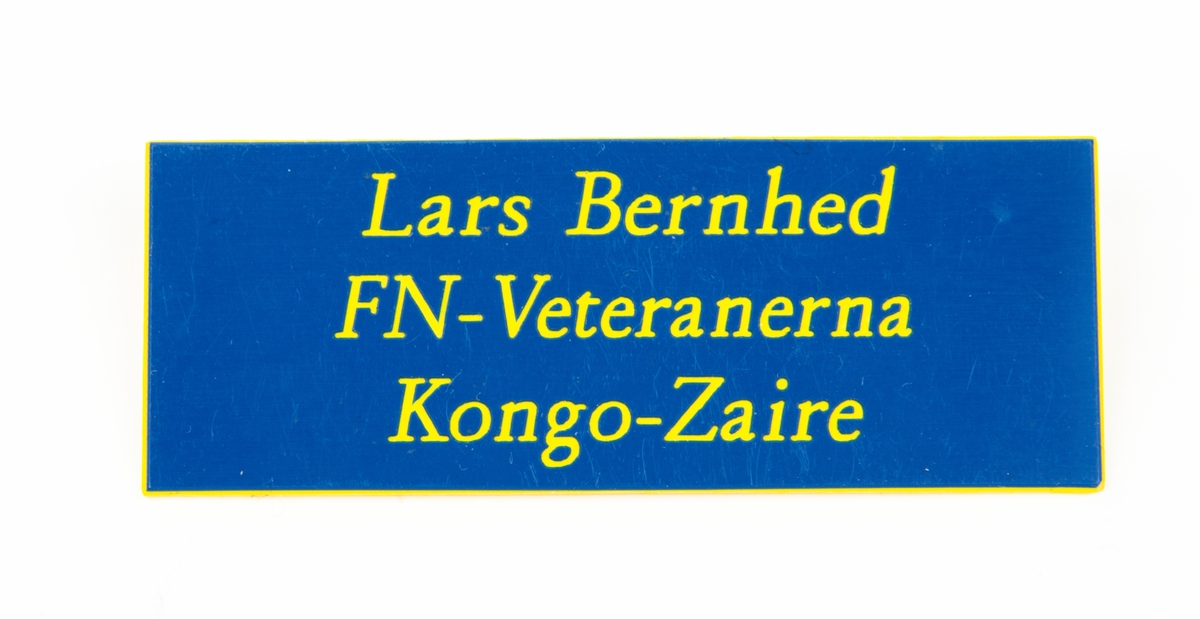 Namnskylt av plast med gul text på blå botten. På baksidan ett fastsättningsspänne. 
Text: Lats Bernhed FN-Veteranerna Kongo-Zaire.