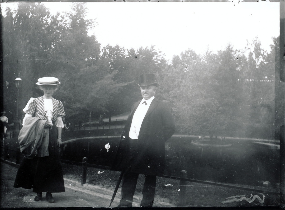 Bestyrer Theodor Rydgren og fru Nicoline Rydgren står selskapskledd i en park. Dam med svane og liten øy i bakgrunnen.