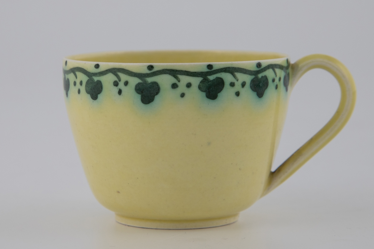 Kaffekopp med tilhørende skål i gulfarget glasur, dekorert med emaljemaling. Dekoren er mørkegrønn, og er utformet som et stilisert planteornament som slynger seg langs fatets og koppens kant.