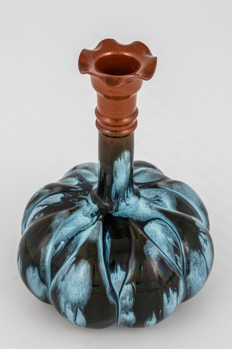 Flaskeformet vase i glasert leirgods. Flattrykket kuleformet korpus med organisk profilering, dekorert med med blå-hvit og sortfarget renneglasur. Høyreist sylinderformet hals, som ender i en utbrettet tunget munningsrand.