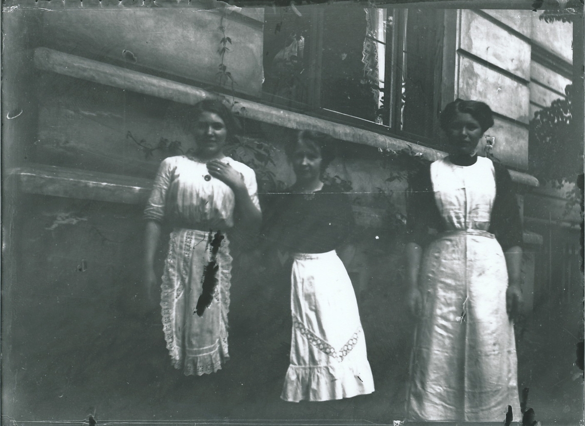 Tre unge kvinner i lange kjoler, med forklær over, står foran bygård i stein. Klatreplanter vokser på bygården.