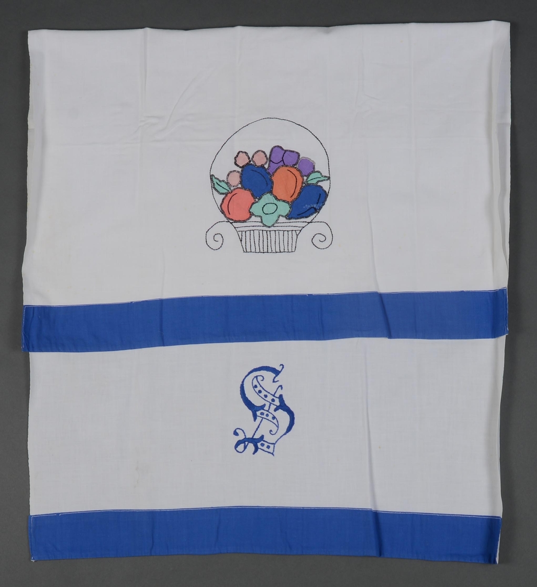 Pyntehåndkle i hvit bomull kantet med blå bomull. Brodert bokstaven "S" ved den ene enden og applikert frukt i flere farger i en brodert kurv. Applikasjonen er kantet med tungesting.