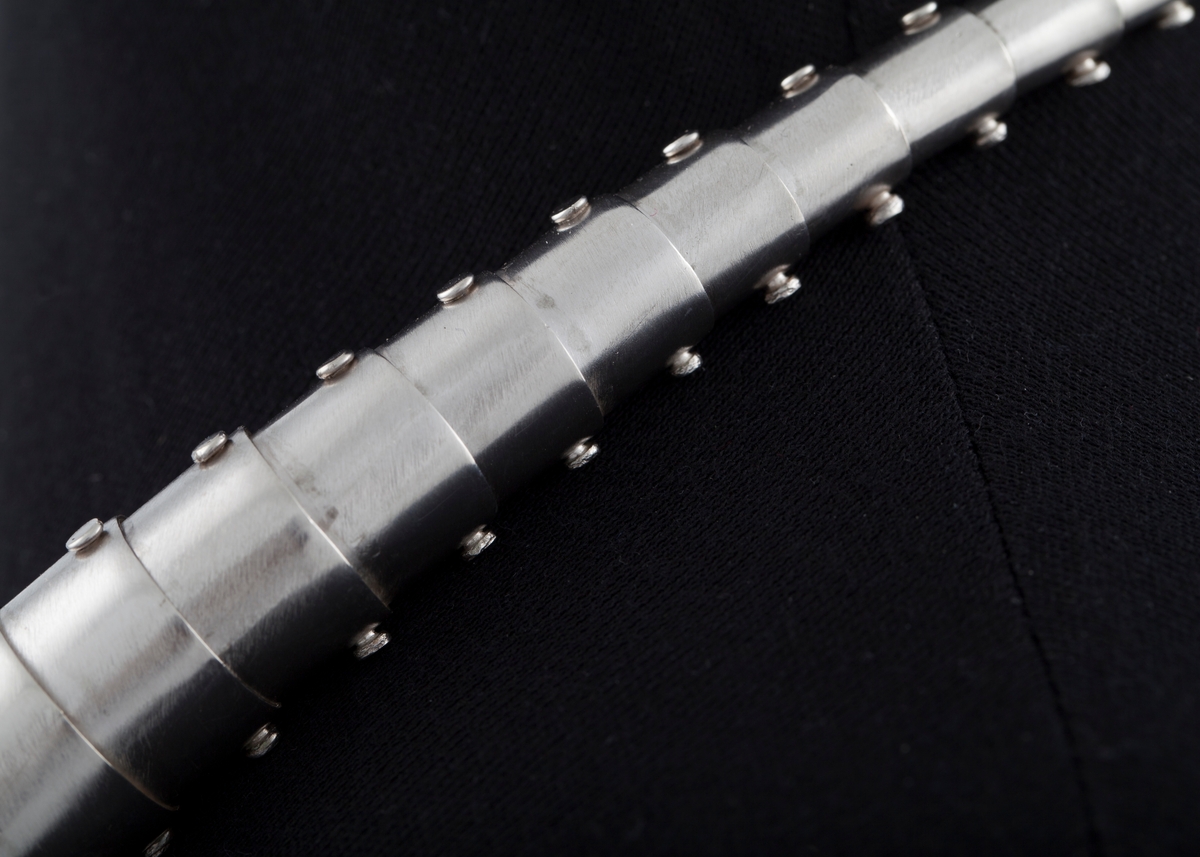 Slangeformet halssmykke i sølv bestående av 55 bevegelige ledd som er tredd i hverandre og minker i størrelse fra midt foran og mot låsen i nakken. Smykket har en symmetrisk form.