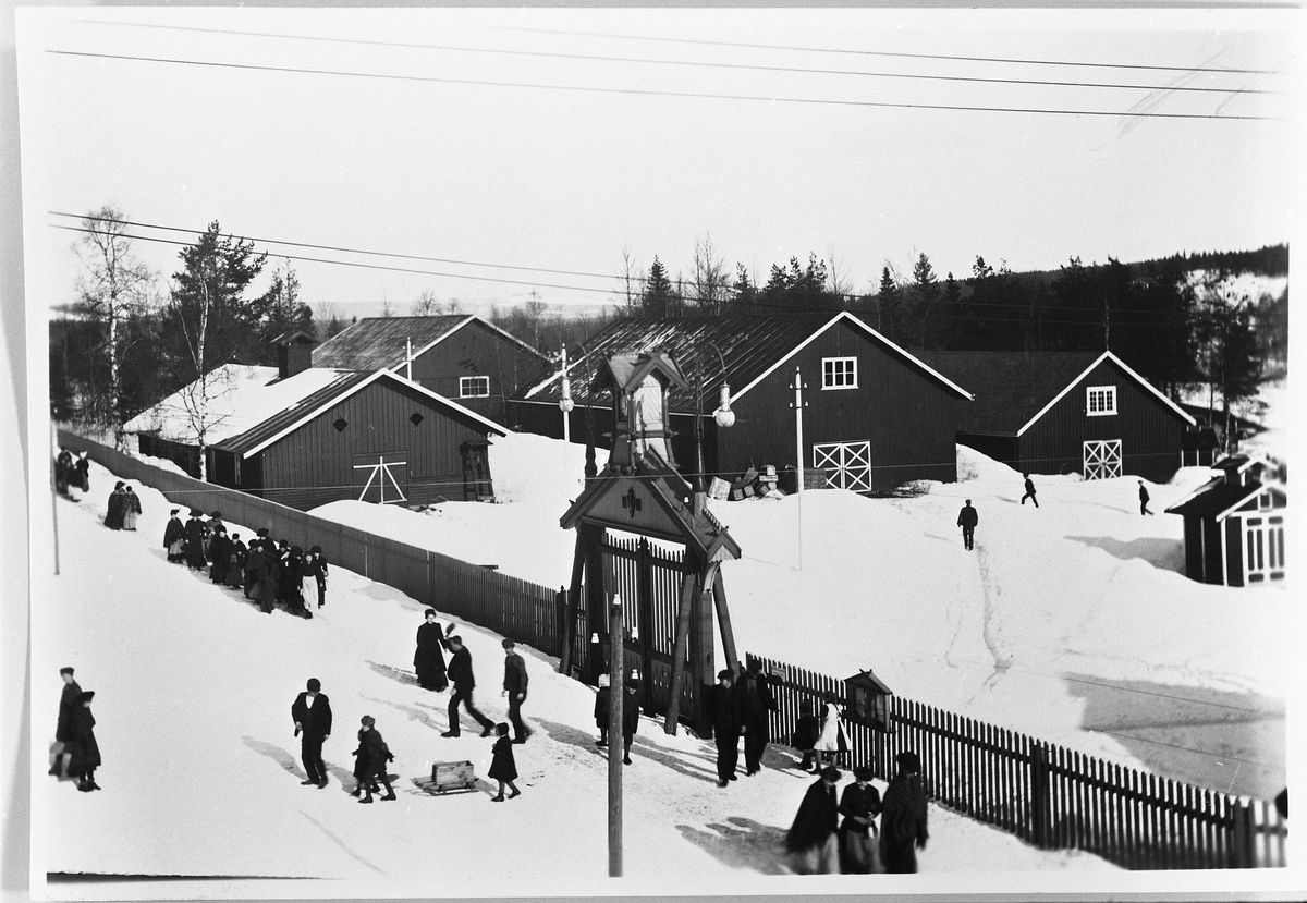 Raufoss Patronfabrikk ca. 1910. Midt i bildet porten inn til fabrikkområdet. Synes å være i middagspausen en vinterdag. Arbeiderne haster ut og inn. Det var middagspause fra 11.30 til kl. 13.00. Husene er magasiner, og et lite brannhus til høgre.