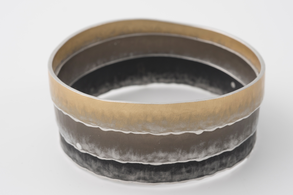 Armbånd bestående av tre flathamrede ringer som er lagt utenpå hverandre og festet sammen med små, blanke nagler. Ringenes ene side er svakt bølgeformet og aluminiumsfarget. Ringenes farger er mørk sotfarget, brun og lys gylden.