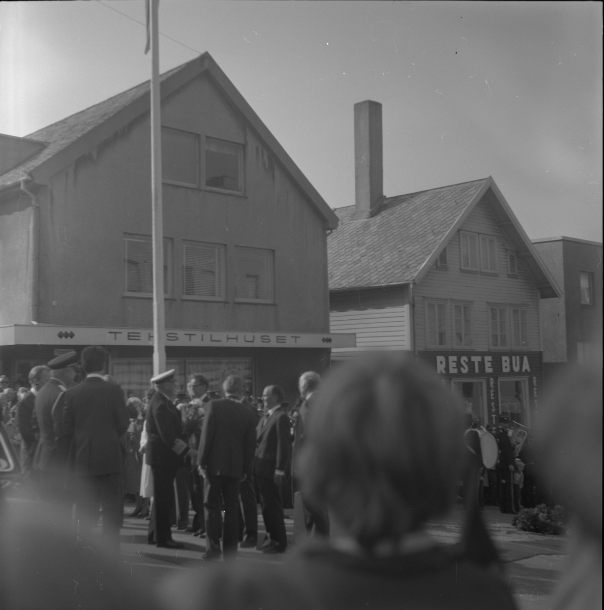 Krysset Haraldsgata/Torggata i Haugesund, 1976. Folkemengd samla i samband med kong Olav V si vitjing i byen. Hans Majestet Kong Olav V til venstre i biletet, med kvit høglue.