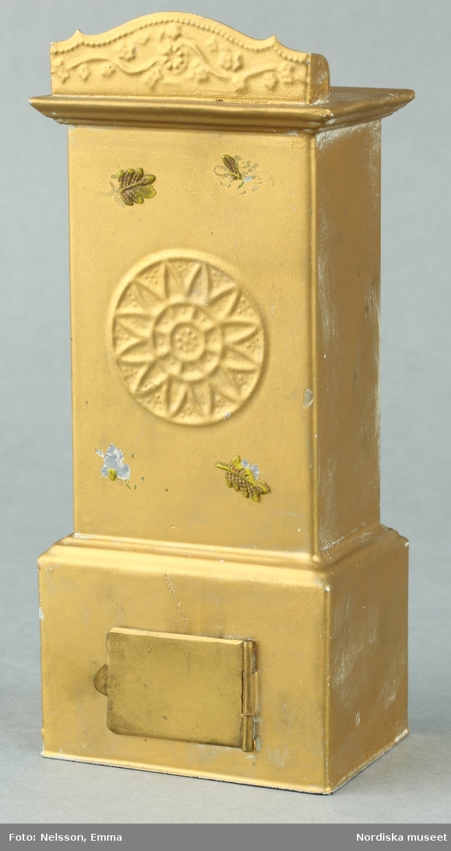 Inventering Sesam 1996-1999:
L 7,3 cm
B 5 cm
H 17,1 cm
Kakelugn till dockskåp, av plåt, bronserad, fyrkantig, sockelparti med eldstadslucka, övre del med blomma i relief (i plåten) och två små påklistrade löv av massa (ursprungligen 4 st), upptill krön med dekor i relief.
Kakelugnen har ursprungligen varit vit (imiterande kakel) vilket fortfarande syns på baksidan.
Tillhör dockskåp inv 164.570. Brukad av givarinnan född 1886.
Leif Wallin nov 1997