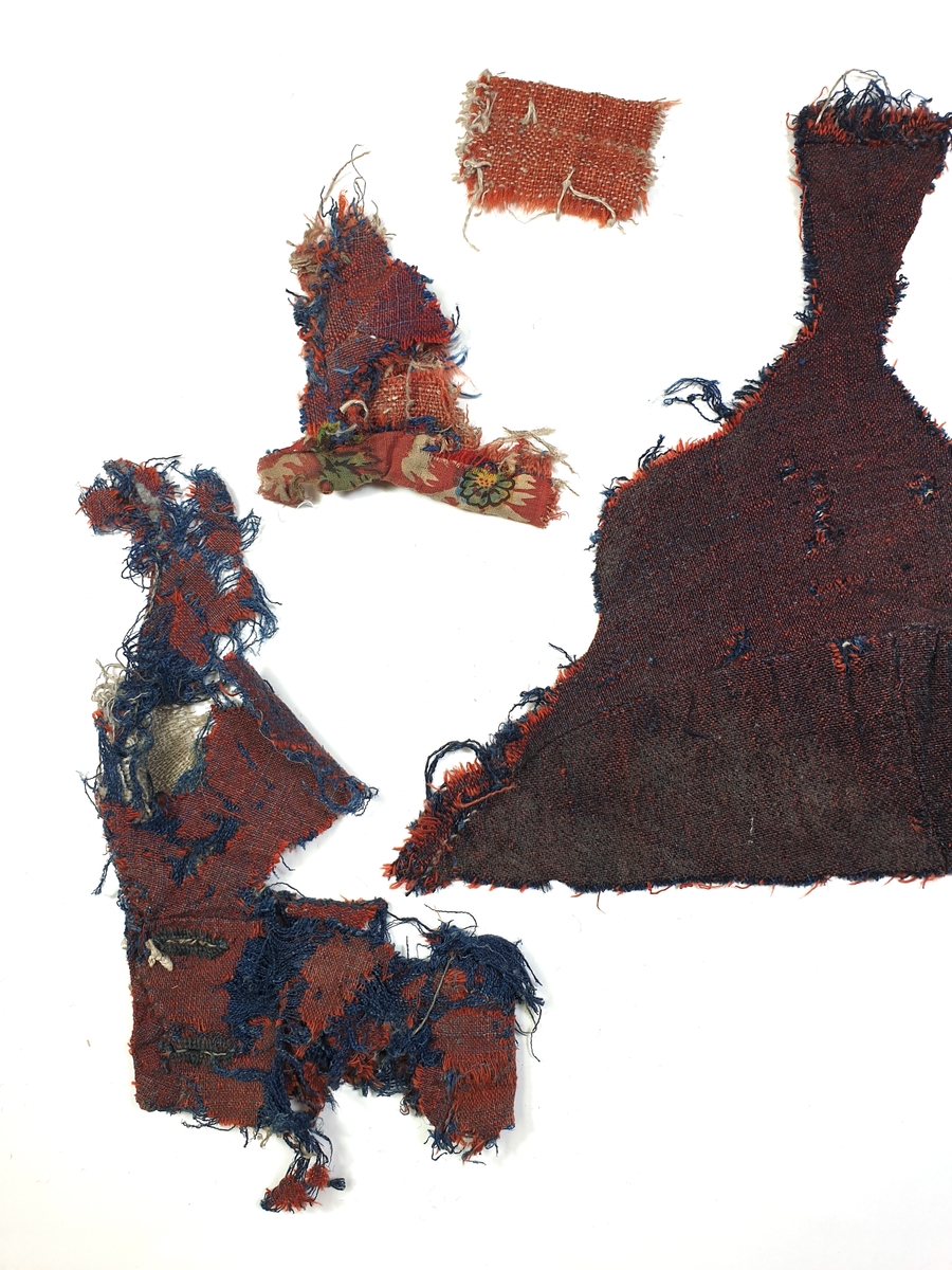Tuskaftat ylletyg, fyra fragment. Blå linvarp, rött ullinslag.
Del av vänster axelstycke. Del av höger ärmslag. Fragment med sömmar och kattuntyg. Lagningstyg.
