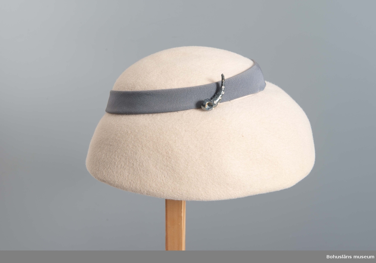 Närmast halvsfärisk hatt i beige filt med svagt antydd kulle kring vilken fästs blågrått ripsband.