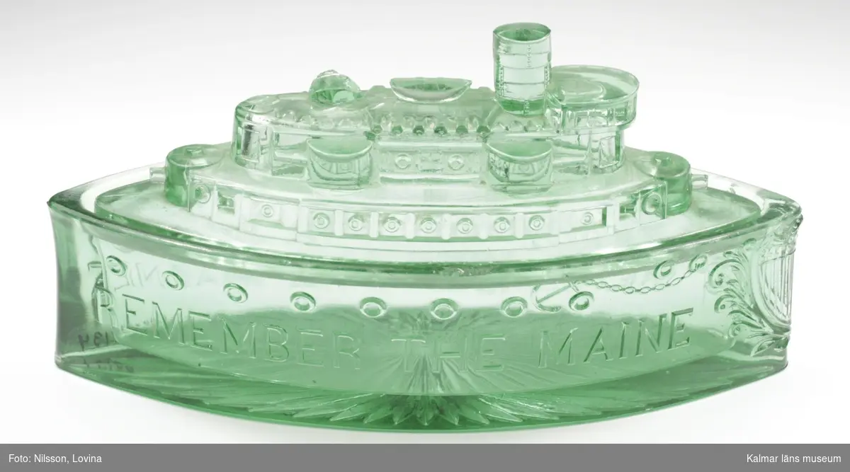 KLM 24134 Ask med lock, av glas. Pressat grönt glas i form av en pansarbåt. Text på långsidorna: Remember the Maine.