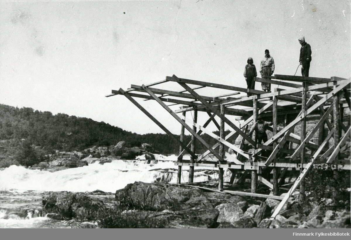 Her ser vi Neiden bru over Neidenelva, under bygging, 1938. Fra venstre ser vi en ukjent mann, Ole Stormo og Wilhelm Kling. Anlegg: Hesseng-Neiden.