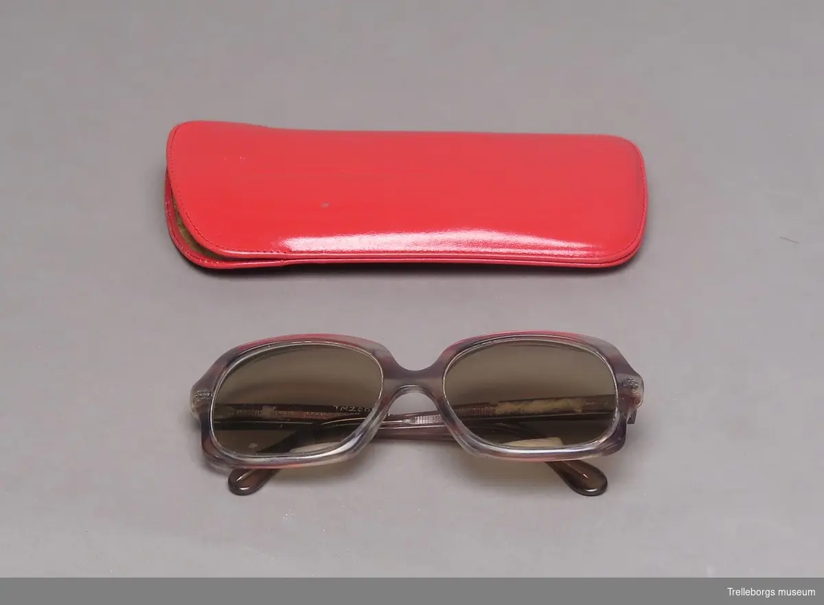 Ett par glasögon med grå-gula plastbågar, med tonade glas. 
Ligger i rött fodral.