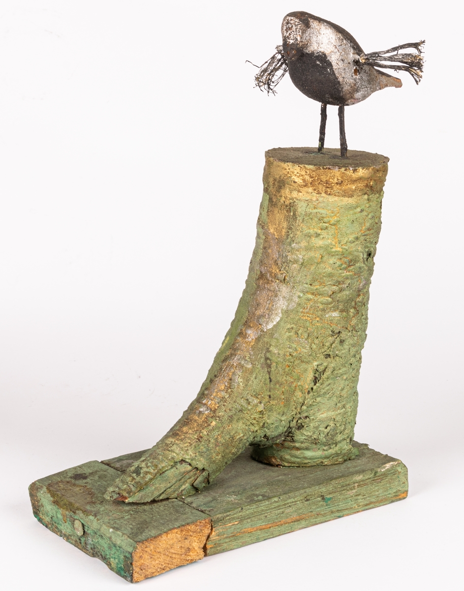 Skulptur av trä. Skulpterad/snidad fågel placerad på en fotliknande sockel, vilken är skapad av en grenklyka. "Foten" är grönmålad, fågeln målad randig i svart och silver, med utbredda vingar av (troligen) tagel.