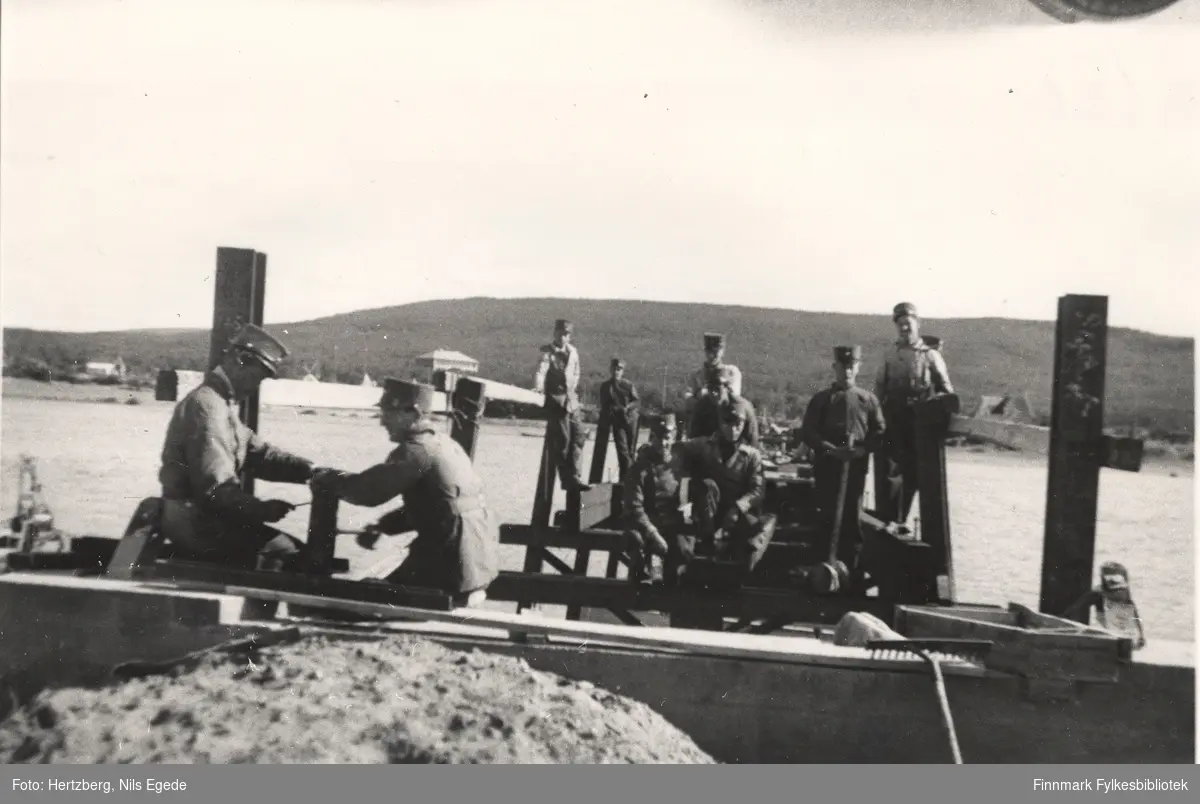 Ti menn holder på med landfeste til bruen over Tanaelven i Seida, 1939. To menn jobber mens resten står eller sitter i bakgrunnen. På den andre siden av elven kan man se noen bygninger.
