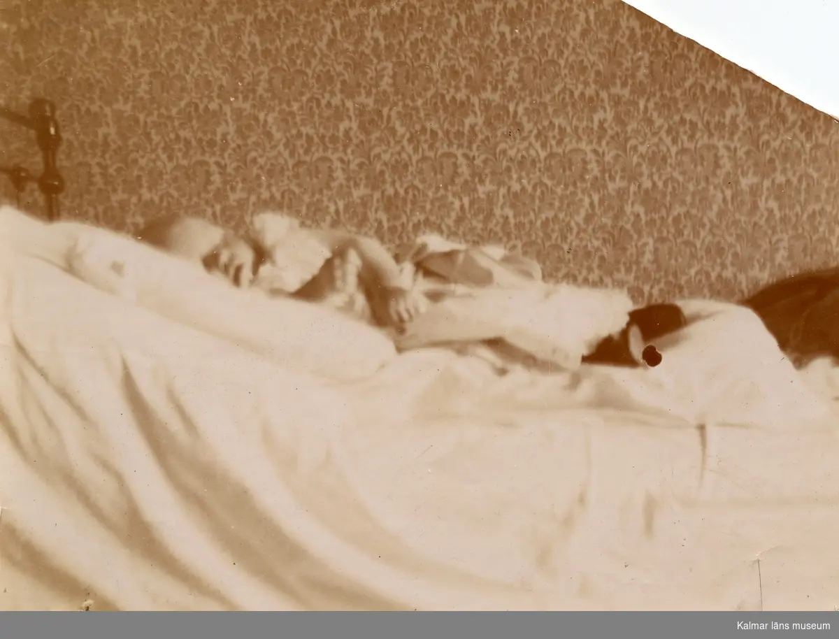 På en säng i ett rum med en blommig tapet ligger ett litet barn och sover. [Curt Nyström Stoopendaal?]