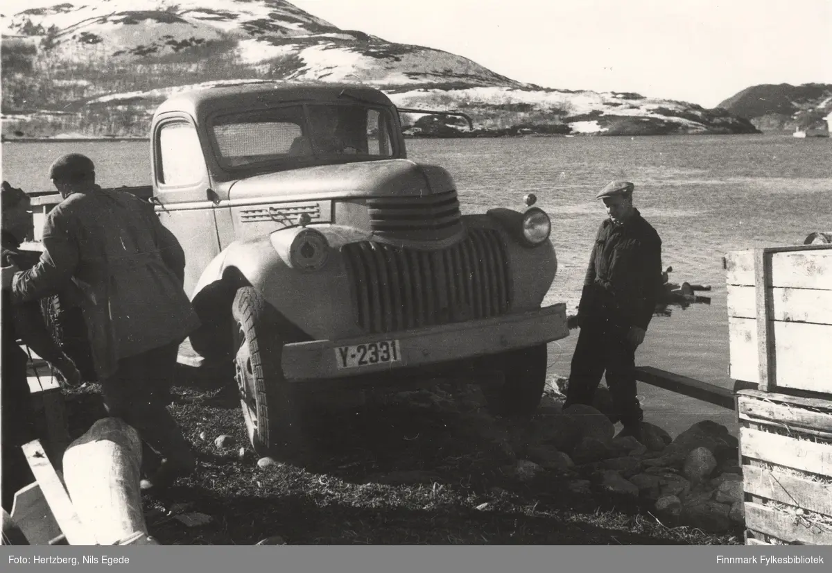 En lastebil (Chevrolet 1946) og noen brubjelker ble losset fra D/S "Slettnes" på en flåte for å frakte det til land, Ifjord-Lebesby 1946. Her kjører lastebilen fra flåten, opp på land. Tre menn, oppsynsmann Johannes Foslund til høyre, ser til at alt går etter planen. Lastebilen y-2331, eier Gustav Simonsen, ble hentet i Smalfjord.