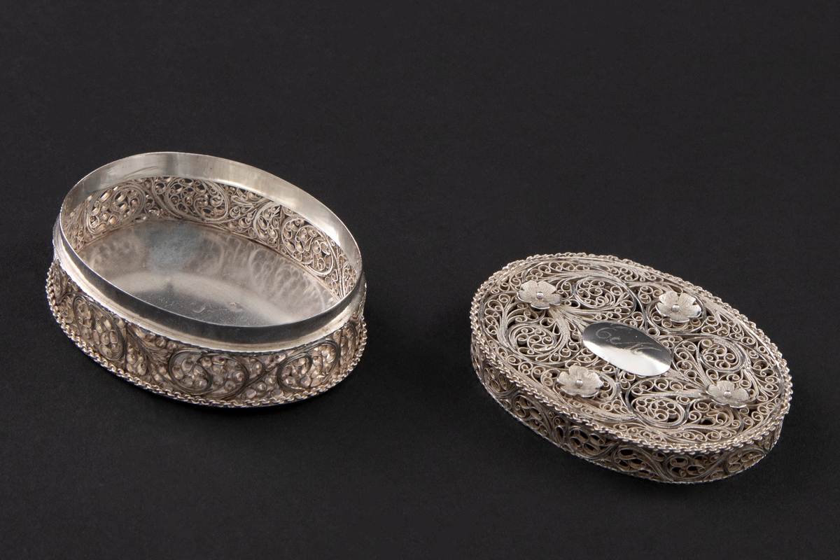 Oval eske av sølvfiligran med lokk. På lokkket er fire små pånittede rosetter rundt en oval, glatt midtplate med initialer.