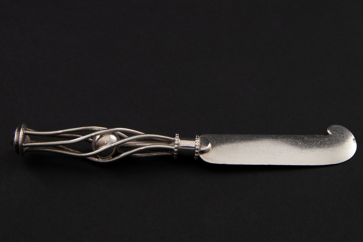 Smørkniv i sølv med tykt, kort knivblad med buet avslutning. Skaftet består av en kule som er innesluttet mellom fem sølvtråder i spiralform. De løper fra et sylinderformet feste ved knivbladet, mot en innfattet slepen ametyst.
