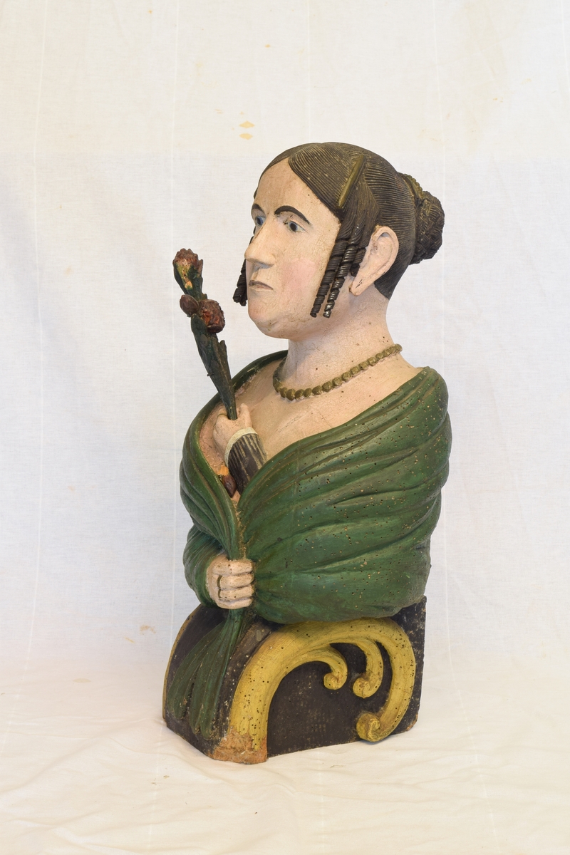 Portrettbyste av kvinnehode. Skulle brukes som gallionsfigurer men kan og ha vært brukt som dekorasjon i et hus. Sortmalt hår med hengekrøller ved ørene og grønt sjal. Holder et sortmalt blad i hånden.