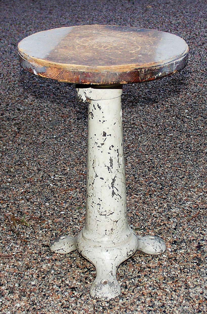 Rund träsits med påspikad masonitskiva. Cylindriskt ben av gråmålat gjutjärn på tre fötter. Bruks- och färgslitage. Proveniens Eiser AB, Borås.