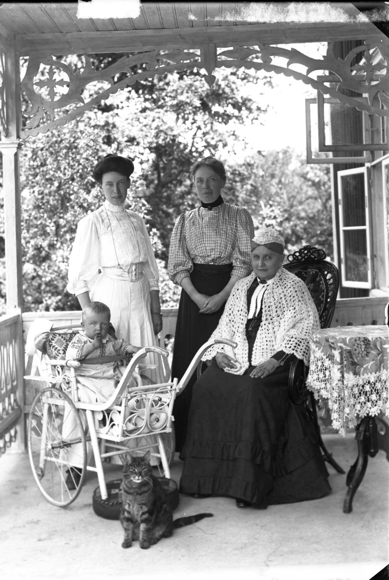 Tre kvinnor, ett barn i en barnvagn och en katt, på en veranda alternativ farstu i Bunn. Den äldre kvinnan, troligen Gunilla Löwenadler, gift med major Löfgren, sitter ner. Den yngre kvinnan i ljust, till vänster, är möjligen deras dotter Gurli Löfgren.