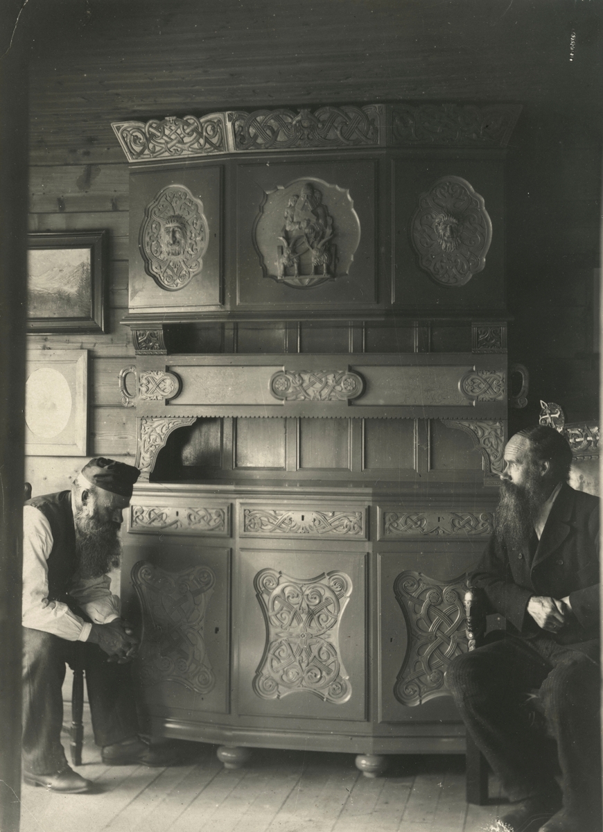 Motiv av to menn som sitter ved et framskap i ei stue. Skapet er dekorert med mytologiske utskjæringer som Tor med hammeren.